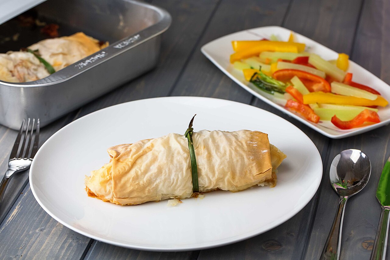 Thin, flaky phyllo pastry sheet wrap with chicken breast peppers olive oil basil goat cheese and sun dried tomatoes on a wood table with stainless steel spoon fork and knife (15785175253)